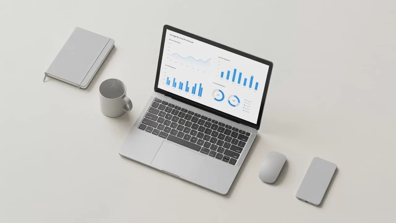 A top-down view of a sleek desk displays a laptop screen showing digital charts and performance metrics. Blue accents highlight specific data trends within a clean, neutral professional workspace setup.