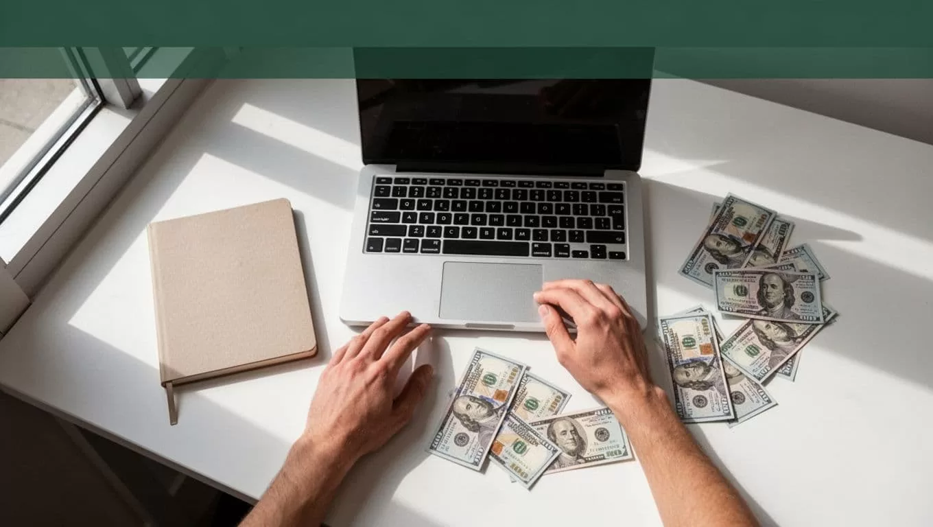 Overhead desk with a laptop, notebook, currency notes, and one person.
