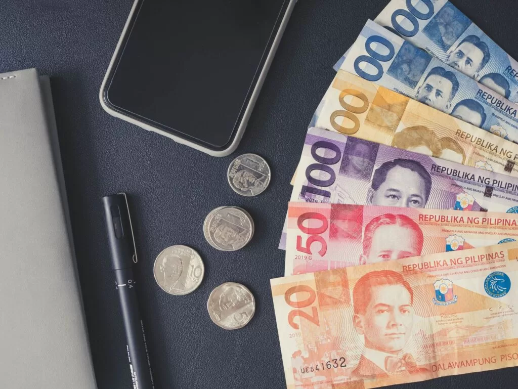 Flat lay of Philippine peso bills, coins, smartphone, and notebook.